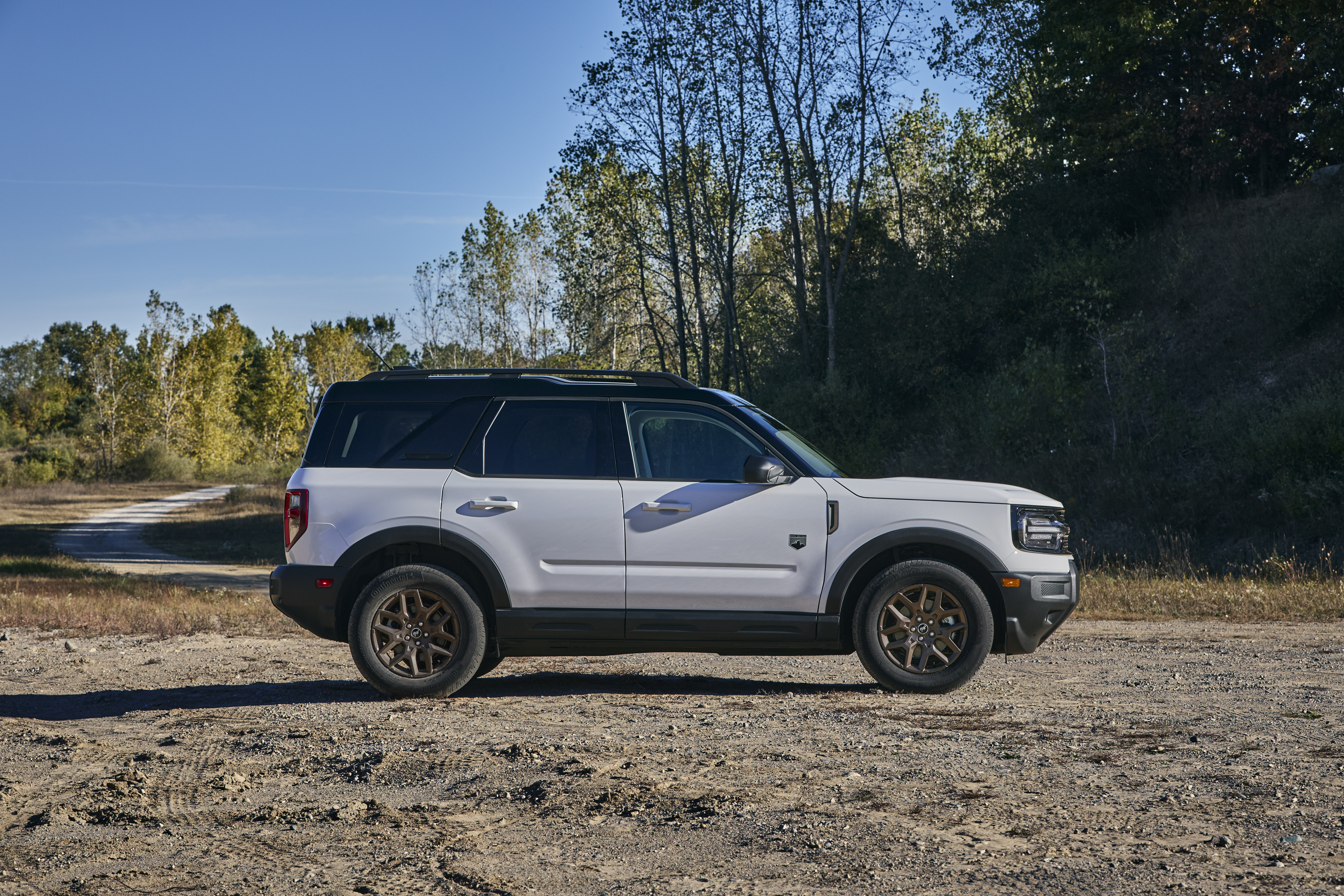 A white Bronco sport with a black top is parked on a gravel lot in a wooded area